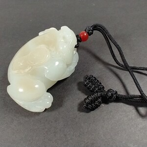 May include: A light-colored jade frog pendant with a black cord and red bead. The frog is carved in a detailed, stylized form. The cord is knotted at the end. The pendant is likely for a necklace.