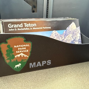 May include: A brown map holder with the National Park Service logo and the text "MAPS". The holder is filled with maps of Grand Teton National Park, John D. Rockefeller, Jr. Memorial Parkway.