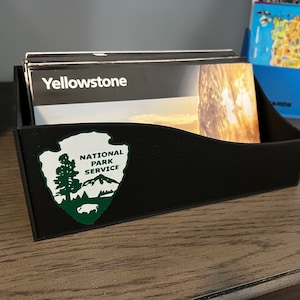 May include: Black storage box with a green National Park Service logo and the word "Yellowstone" on a stack of brochures inside.