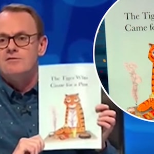 Op de afbeelding: Een man met een bril houdt een boek vast met de titel "The Tiger Who Came for a Pint". De omslag van het boek toont een oranje tijger met zwarte strepen, zittend aan een tafel met een pint bier. De achtergrond is een blauwe studio.
