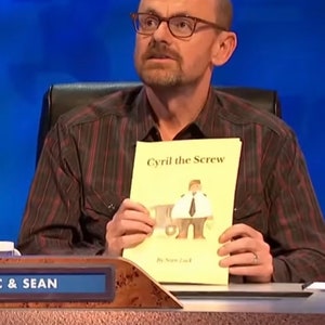 Peut inclure: Un homme portant des lunettes et une chemise à rayures marron tient un livre intitulé "Cyril the Screw" avec une illustration d'une personne en uniforme. Le livre est de Sean Lock.