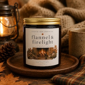 May include: A dark glass candle jar with a gold lid and a cream-colored label. The label reads Coastal Oak Candle Co., flannel & firelight, vanilla, cinnamon, warm musk, and coconut beeswax, 7.3 oz, 207 g, with a fireplace illustration.