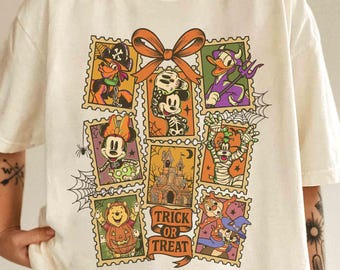 May include: Cream-colored t-shirt featuring a Halloween-themed graphic. The graphic includes postage stamp-style images of cartoon characters, a castle, and the words "Trick or Treat." A large orange bow is at the top of the design.