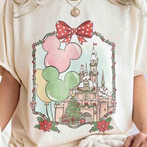 K&ouml;nnte beinhalten: Cremefarbenes T-Shirt mit festlichem Aufdruck. Das Design zeigt Mickey Mouse-f&ouml;rmige Ballons in Rosa, Gr&uuml;n und Gelb, eine rote Schleife mit Punkten und eine Schlossszene mit einem Weihnachtsbaum. Das Shirt hat eine lockere Passform.