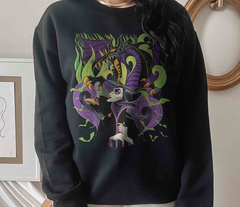 May include: A black crewneck sweatshirt featuring a graphic print of a purple and black dragon looming over a character with pale skin and a black headpiece, accompanied by a black bird and green flames, with small green bats scattered throughout.