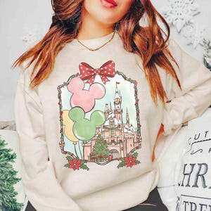 K&ouml;nnte beinhalten: Cremefarbenes Sweatshirt mit festlichem Disney-Schloss-Design. Die Grafik zeigt Mickey Mouse-Ballons in Rosa, Gelb und Gr&uuml;n, eine rote Schleife mit Punkten und einen Weihnachtsbaum. Das Design ist mit einem dekorativen Rahmen versehen.