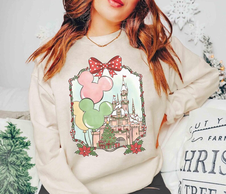 K&ouml;nnte beinhalten: Cremefarbenes Sweatshirt mit festlichem Disney-Schloss-Design. Die Grafik zeigt Mickey Mouse-Ballons in Rosa, Gelb und Gr&uuml;n, eine rote Schleife mit Punkten und einen Weihnachtsbaum. Das Design ist mit einem dekorativen Rahmen versehen.