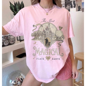 May include: A light pink t-shirt with a graphic that reads "The Most Magical Place on Earth." The graphic features a castle, a silhouette of a person and child, and other park elements. The shirt has short sleeves.