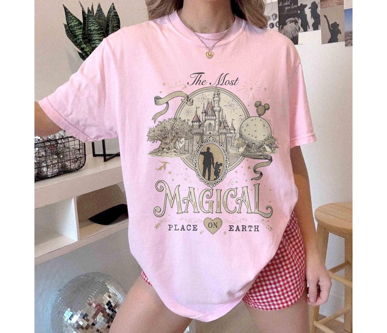 May include: A light pink t-shirt with a graphic that reads "The Most Magical Place on Earth." The graphic features a castle, a silhouette of a person and child, and other park elements. The shirt has short sleeves.
