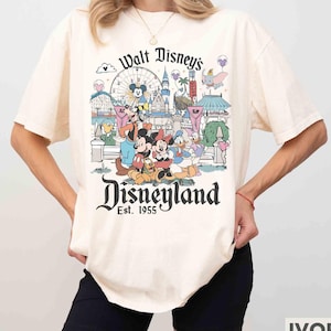 May include: Ivory-colored t-shirt featuring a colorful Disneyland graphic. The design includes Mickey and Minnie Mouse, Donald Duck, and other Disney characters in front of a Ferris wheel and castle. The text "Walt Disney's Disneyland" and "Est. 1955" are also visible.