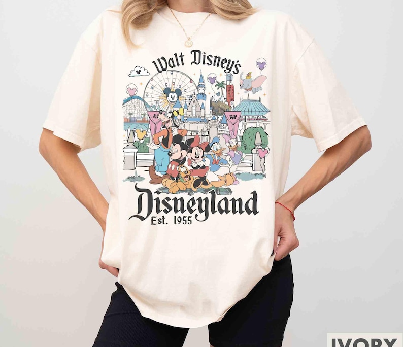 May include: Ivory-colored t-shirt featuring a colorful Disneyland graphic. The design includes Mickey and Minnie Mouse, Donald Duck, and other Disney characters in front of a Ferris wheel and castle. The text "Walt Disney's Disneyland" and "Est. 1955" are also visible.