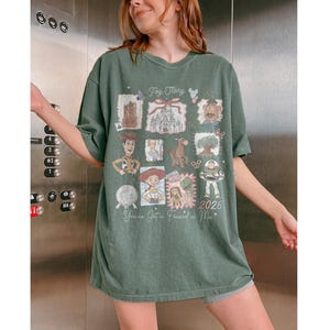 以下が含まれることがあります： トイ・ストーリーのコラージュデザインが施されたグリーンのTシャツ。映画の様々なキャラクターや画像、テキスト「Toy Story」、「You've Got a Friend in Me」、そして年号「2026」が特徴です。このTシャツはオーバーサイズです。