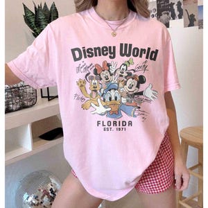 May include: A light pink t-shirt with the words "Disney World" in black, featuring cartoon characters like Mickey Mouse, Minnie Mouse, and Goofy. The shirt also has the text "Florida EST. 1971".