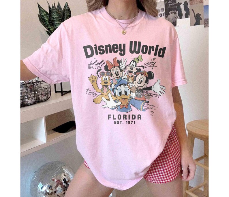 May include: A light pink t-shirt with the words "Disney World" in black, featuring cartoon characters like Mickey Mouse, Minnie Mouse, and Goofy. The shirt also has the text "Florida EST. 1971".