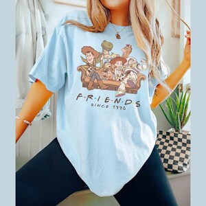 Może przedstawiać: Jasnoniebieska koszulka z nadrukiem graficznym postaci z Toy Story siedzących na kanapie. Pod obrazkiem znajduje się napis "F.R.I.E.N.D.S. SINCE 1995".