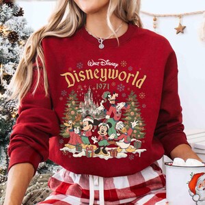 May include: A red sweatshirt featuring a Disney World graphic with Mickey Mouse, Minnie Mouse, Donald Duck, Goofy, and other characters. The graphic includes the text "Walt Disney Disneyworld 1971" and Christmas trees.