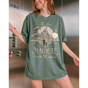 May include: A green t-shirt with the text "The Most Magical Place on Earth" in a vintage style. The graphic includes a castle, a tree, and a silhouette of a person. The shirt is oversized and has short sleeves.