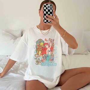 May include: White t-shirt featuring a graphic print of Ariel, the Little Mermaid, with Flounder and Sebastian. The text "The Little Mermaid" is printed below the image.