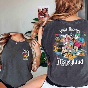 以下が含まれることがあります： ヴィンテージ風のグレーのTシャツ2枚。1枚はミッキーマウスのグラフィックと「Daddy」という文字が書かれている。もう1枚はミッキーマウス、ドナルドダック、グーフィー、プルート、その他のディズニーキャラクターのグラフィックと「Walt Disney's Disneyland Est. 1955」という文字が書かれている。