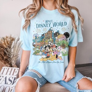 May include: Pale blue t-shirt with a graphic of Disney characters and attractions, including the text "Walt Disney World" and "The Most Magical Place on Earth". The shirt has a crew neck and short sleeves.