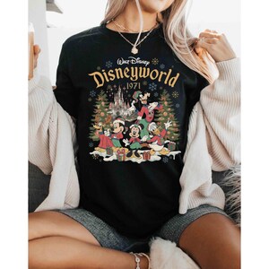May include: Black crew neck t-shirt featuring a colorful graphic of Disney characters in front of a castle and Christmas trees. The text "Walt Disney Disneyworld 1971" is printed in gold across the top of the graphic.