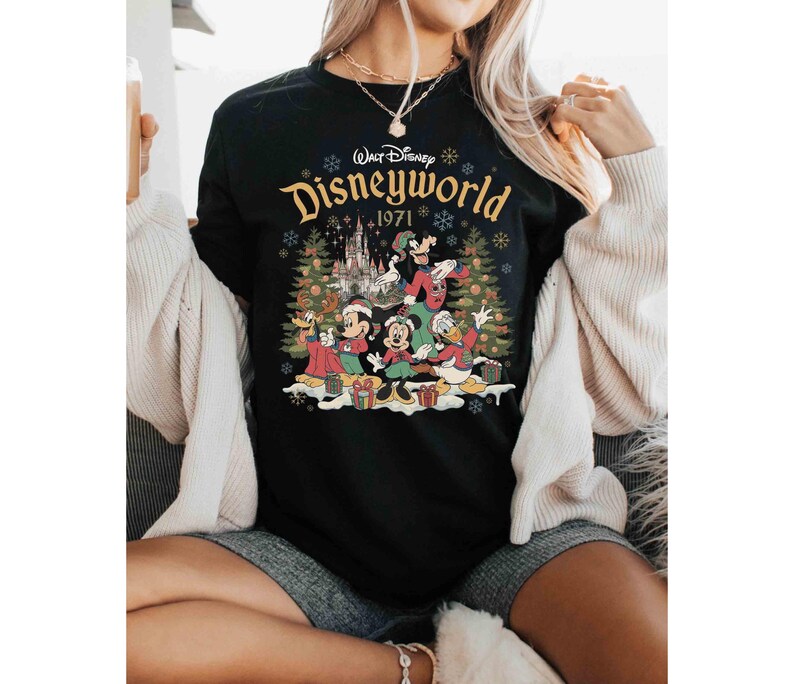 May include: Black crew neck t-shirt featuring a colorful graphic of Disney characters in front of a castle and Christmas trees. The text "Walt Disney Disneyworld 1971" is printed in gold across the top of the graphic.