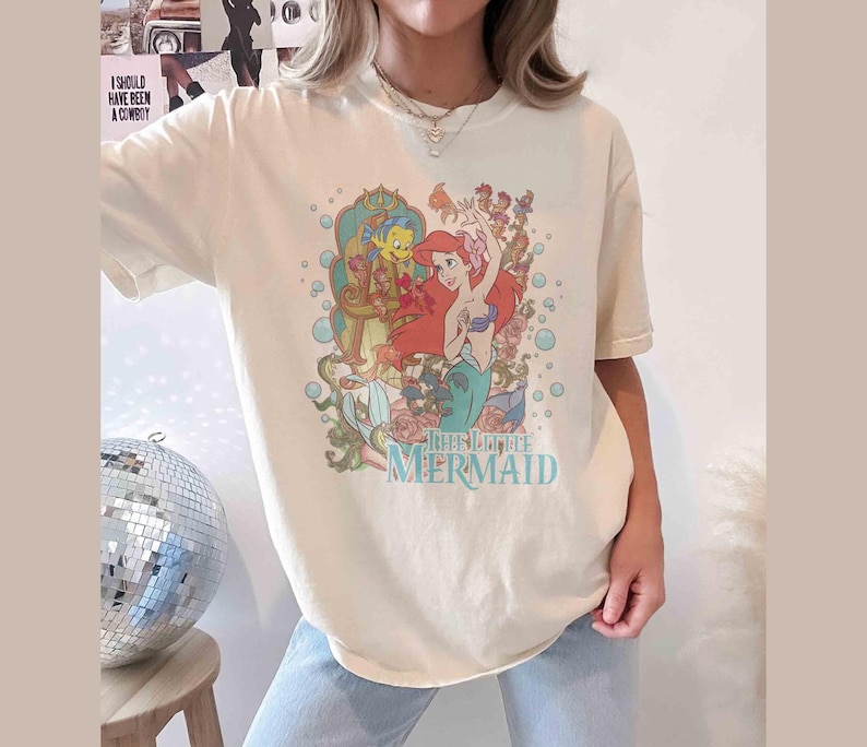 May include: A white t-shirt featuring a graphic print of Ariel from The Little Mermaid. The graphic depicts Ariel with her red hair and tail, surrounded by bubbles and sea creatures. The text "The Little Mermaid" is printed beneath the graphic.