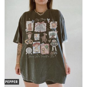 以下が含まれることがあります： トイ・ストーリーのデザインが施された濃い緑色のTシャツ。シャツには、さまざまなキャラクターのイラストと「Toy Story」、「You've Got a Friend in Me」、そして年号「2026」の文字がプリントされています。リラックスフィットのTシャツにデザインがプリントされています。