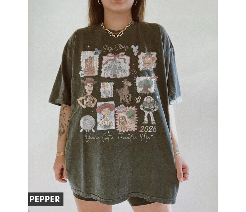 以下が含まれることがあります： トイ・ストーリーのデザインが施された濃い緑色のTシャツ。シャツには、さまざまなキャラクターのイラストと「Toy Story」、「You've Got a Friend in Me」、そして年号「2026」の文字がプリントされています。リラックスフィットのTシャツにデザインがプリントされています。