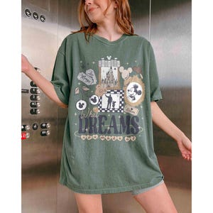 K&ouml;nnte beinhalten: Gr&uuml;nes T-Shirt mit einem Grafikdesign mit Mickey Mouse, einem Schloss und den Worten "What Dreams Are Made Of". Das Design umfasst Mickey Mouse-Ohren, eine Silhouette und dekorative Rahmen. Das Shirt hat eine lockere Passform.