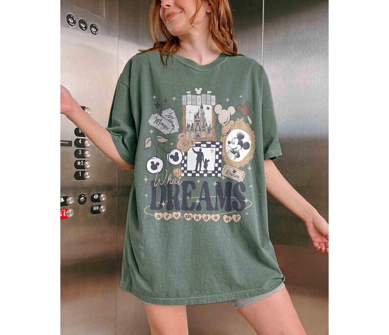 K&ouml;nnte beinhalten: Gr&uuml;nes T-Shirt mit einem Grafikdesign mit Mickey Mouse, einem Schloss und den Worten "What Dreams Are Made Of". Das Design umfasst Mickey Mouse-Ohren, eine Silhouette und dekorative Rahmen. Das Shirt hat eine lockere Passform.