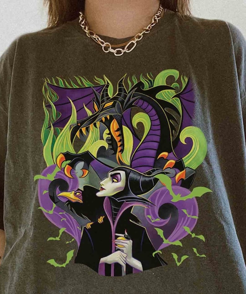 May include: A charcoal gray t-shirt featuring a graphic print of a dark-haired character in a black robe, a large purple and black dragon, a black bird, and green bats. The design includes swirling green and purple smoke elements.