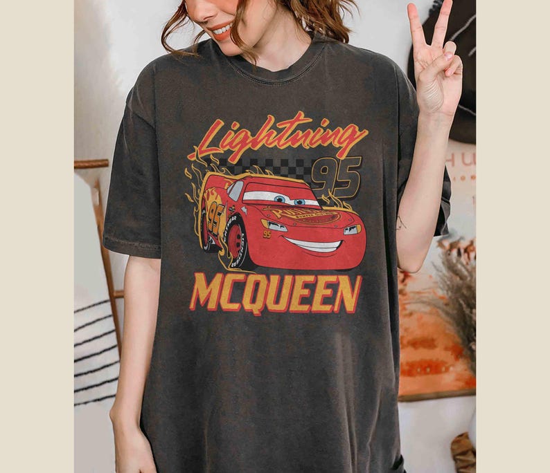 Peut inclure: Un t-shirt noir &agrave; l'aspect vieilli avec une image de dessin anim&eacute; de Flash McQueen du film Cars de Disney Pixar. Le texte "Lightning 95 McQueen" est imprim&eacute; en jaune sur le t-shirt.
