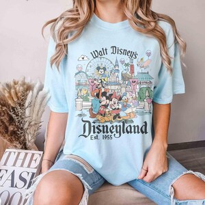 May include: Light blue t-shirt featuring a graphic of Disneyland with Mickey Mouse, Minnie Mouse, and other Disney characters. The shirt has the text "Walt Disney's Disneyland Est. 1955" in a vintage style.