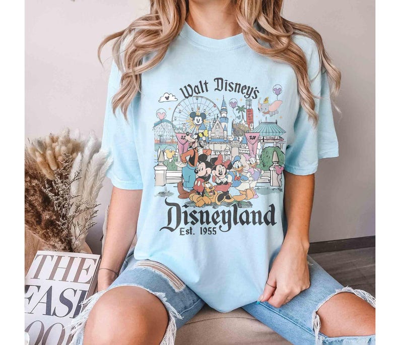 May include: Light blue t-shirt featuring a graphic of Disneyland with Mickey Mouse, Minnie Mouse, and other Disney characters. The shirt has the text "Walt Disney's Disneyland Est. 1955" in a vintage style.