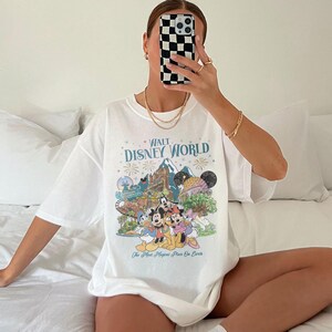 May include: White t-shirt with a colourful graphic of Disney characters and the text "Walt Disney World". The design includes iconic park imagery and the phrase "The Most Magical Place On Earth".