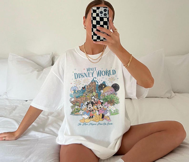 May include: White t-shirt with a colourful graphic of Disney characters and the text "Walt Disney World". The design includes iconic park imagery and the phrase "The Most Magical Place On Earth".