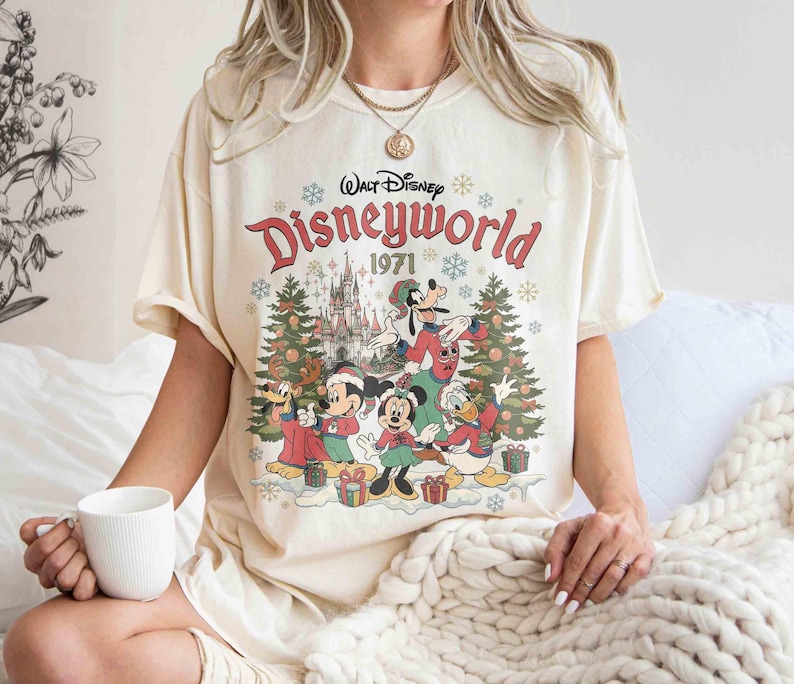 May include: Cream-colored t-shirt featuring a graphic of Disney characters in a winter scene with Christmas trees and gifts. The text "Walt Disney Disneyworld 1971" is printed above the characters. The shirt has short sleeves.