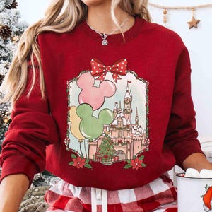 K&ouml;nnte beinhalten: Ein rotes Sweatshirt mit festlichem Design. Die Grafik zeigt Mickey Mouse-f&ouml;rmige Ballons in Rosa, Gr&uuml;n und Gelb, ein Schloss, einen Weihnachtsbaum und eine rote Schleife mit Tupfen. Das Sweatshirt hat eine einfarbige rote Farbe.