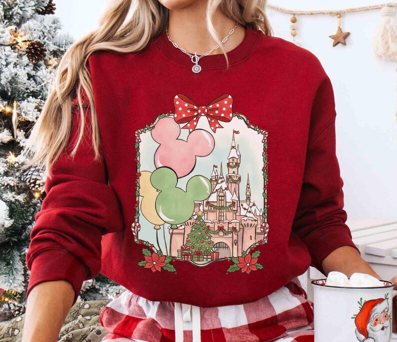 K&ouml;nnte beinhalten: Ein rotes Sweatshirt mit festlichem Design. Die Grafik zeigt Mickey Mouse-f&ouml;rmige Ballons in Rosa, Gr&uuml;n und Gelb, ein Schloss, einen Weihnachtsbaum und eine rote Schleife mit Tupfen. Das Sweatshirt hat eine einfarbige rote Farbe.