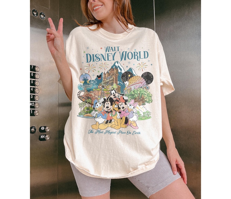 May include: Cream-coloured t-shirt with a colourful graphic of Disney characters and landmarks, featuring the text "WALT DISNEY WORLD" and "The Most Magical Place On Earth". The shirt has a crew neck and short sleeves.