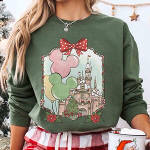 K&ouml;nnte beinhalten: Gr&uuml;nes Sweatshirt mit festlichem Design. Das Design umfasst Mickey Mouse-Ballons, ein Schloss und einen Weihnachtsbaum, umrahmt von einer roten Schleife mit Punkten und Weihnachtssternen. Das Sweatshirt hat eine einfarbige gr&uuml;ne Farbe.