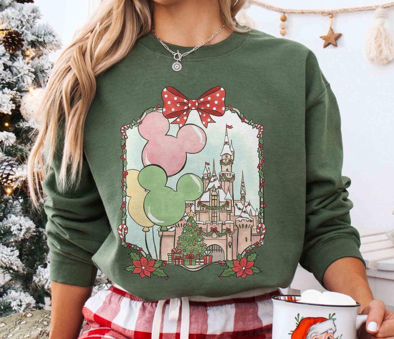 K&ouml;nnte beinhalten: Gr&uuml;nes Sweatshirt mit festlichem Design. Das Design umfasst Mickey Mouse-Ballons, ein Schloss und einen Weihnachtsbaum, umrahmt von einer roten Schleife mit Punkten und Weihnachtssternen. Das Sweatshirt hat eine einfarbige gr&uuml;ne Farbe.