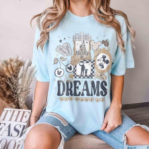 K&ouml;nnte beinhalten: Hellblaues T-Shirt mit einem grafischen Design mit einem Schloss, Mickey Mouse und dem Text "Where Dreams are Made Of". Das Design umfasst Sterne, Kirschen und ein gerahmtes Mickey Mouse-Bild.