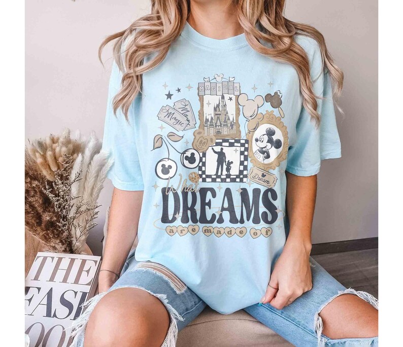 K&ouml;nnte beinhalten: Hellblaues T-Shirt mit einem grafischen Design mit einem Schloss, Mickey Mouse und dem Text "Where Dreams are Made Of". Das Design umfasst Sterne, Kirschen und ein gerahmtes Mickey Mouse-Bild.