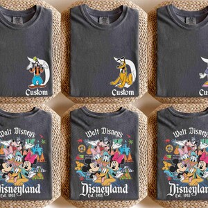 以下が含まれることがあります： 異なるデザインのグレーのTシャツ6枚。最初の3枚のシャツは、プルート、グーフィー、デイジー・ダックなど、ディズニーの漫画のキャラクターが描かれています。最後の3枚のシャツは、ミッキーマウス、ミニーマウス、ドナルドダック、グーフィーなど、ディズニーのキャラクターのグループが描かれています。シャツにはすべて、「Walt Disney's Disneyland Est. 1955」という文字が印刷されています。
