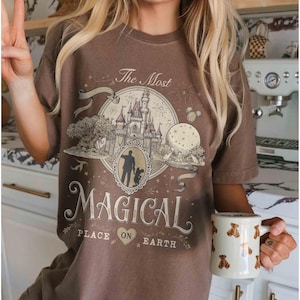 May include: Brown t-shirt with a graphic featuring a castle, a silhouette of a person and child, and the text "The Most Magical Place on Earth." A white mug with bear designs is held in the hand.