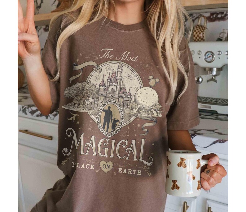 May include: Brown t-shirt with a graphic featuring a castle, a silhouette of a person and child, and the text "The Most Magical Place on Earth." A white mug with bear designs is held in the hand.
