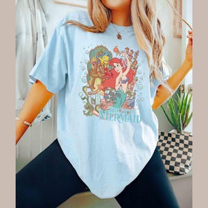 May include: A light blue t-shirt featuring a graphic print of Ariel, the Little Mermaid, and Flounder. The graphic is surrounded by bubbles and flowers. The text "The Little Mermaid" is printed below the graphic.