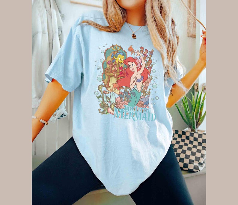 May include: A light blue t-shirt featuring a graphic print of Ariel, the Little Mermaid, and Flounder. The graphic is surrounded by bubbles and flowers. The text "The Little Mermaid" is printed below the graphic.
